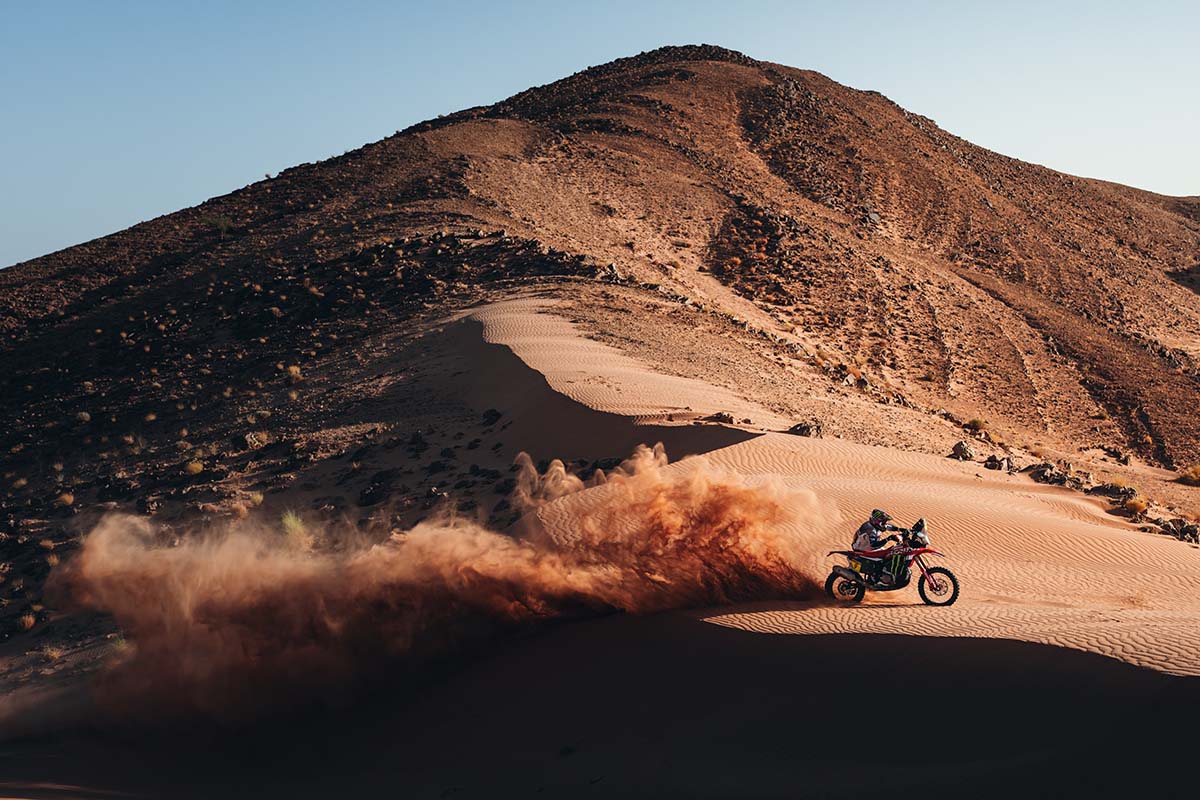 Rally rider on a red Honda motorcycle in a sand dune in front of a rocky mountain, with a cloud of sand behind him