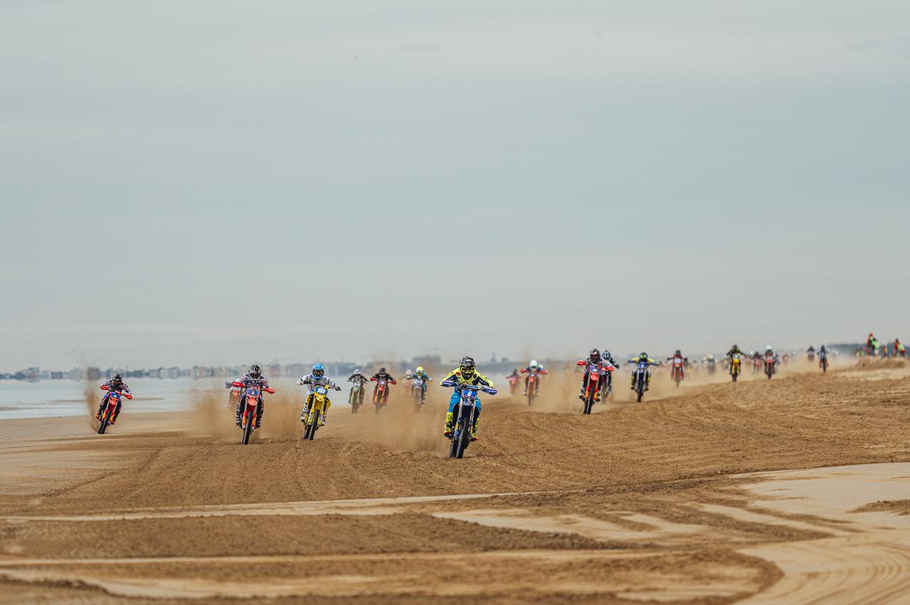 Riders in a sand race cross a beach in a wide pack, with sand spraying up behind them