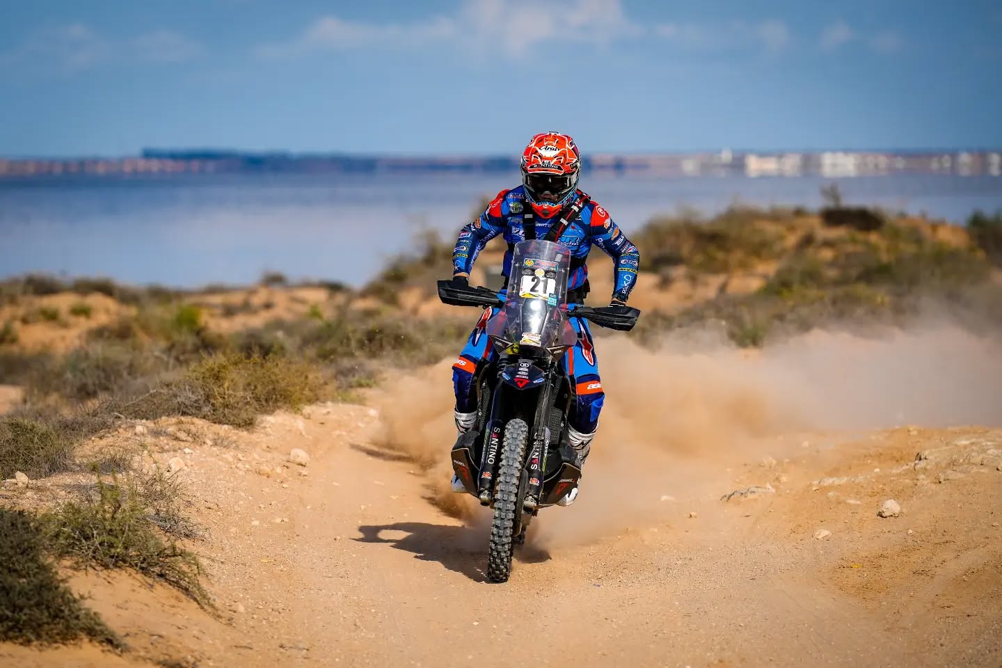 Motorradfahrer mit Startnummer 21 auf einer staubigen Sandpiste, im Hintergrund Wasser