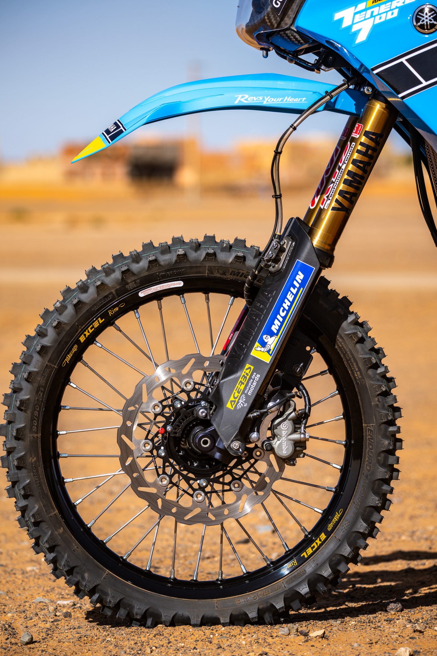 Front wheel of a blue Yamaha T&eacute;n&eacute;r&eacute; 700 with a Michelin enduro tyre, black Excel rim and brake disc against a desert backdrop