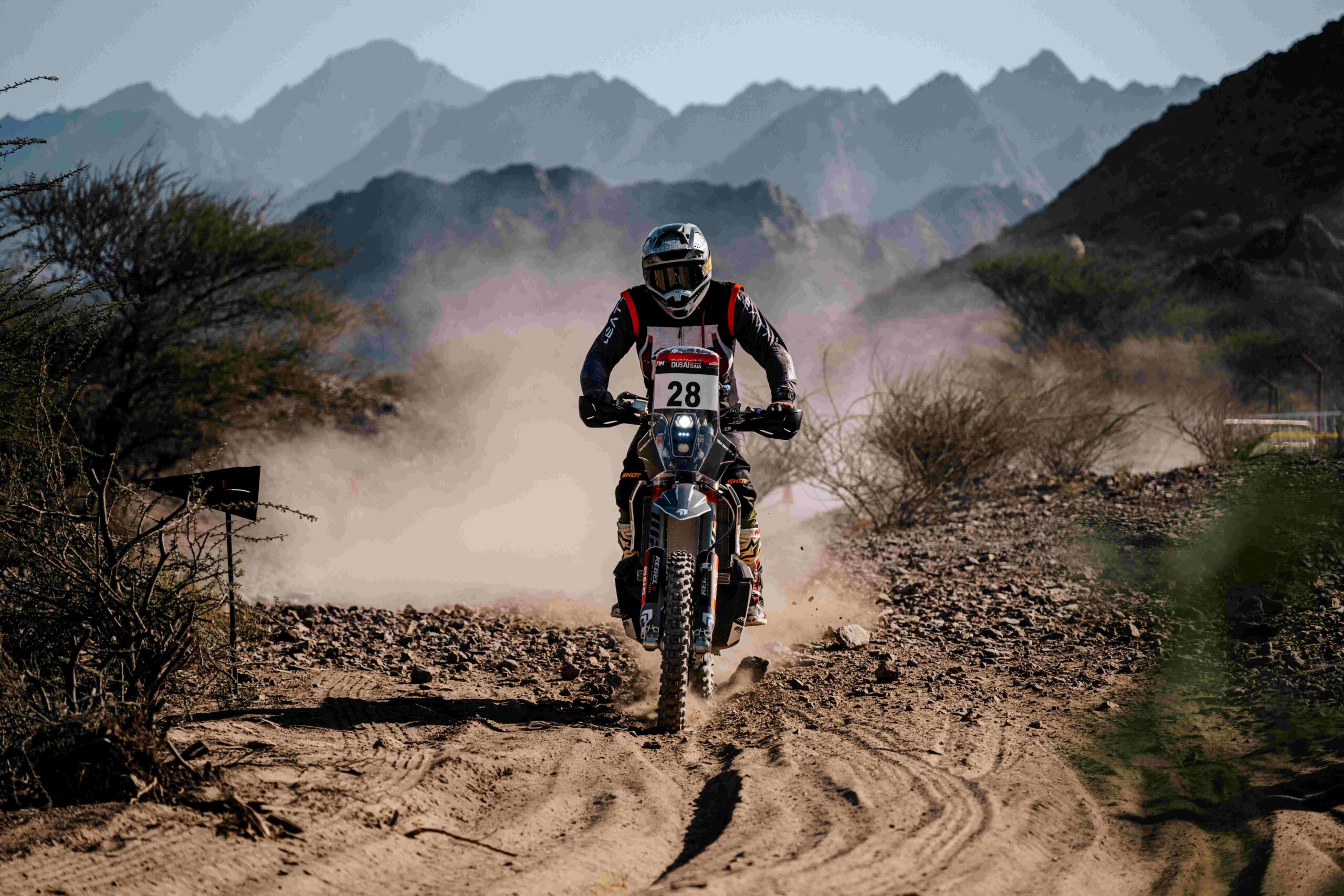 Rally motorcyclist with number 28 riding on a dusty gravel track with mountains in the background