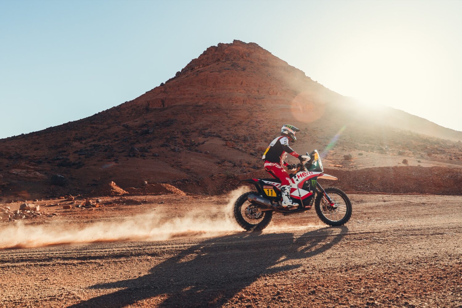 Rally rider on a Hero motorcycle with number 11 kicking up a dust trail in front of a rocky mountain