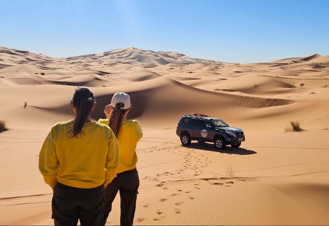 Two women in yellow sweaters look at a dark off-road vehicle in the sand dunes