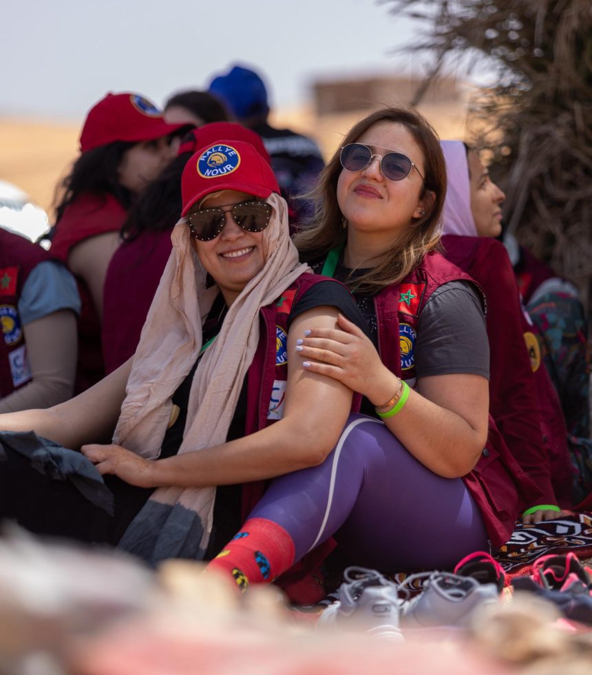 Two smiling participants in red vests and caps reading Rallye Nour sit in a group