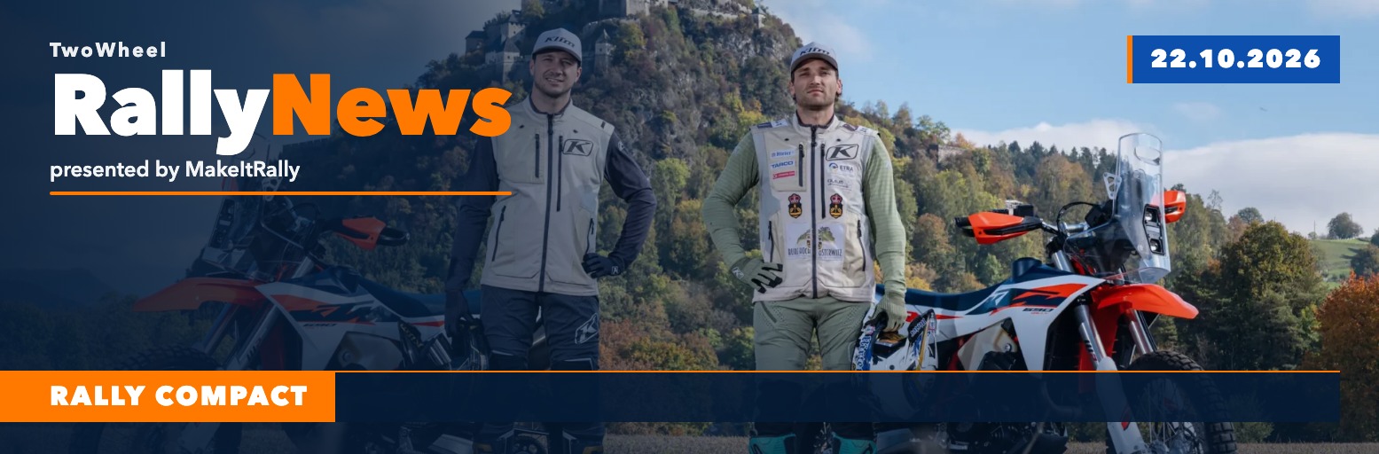 Two rally riders stand next to two KTM 690 Rally bikes in a field, with a castle on a wooded hill in the background.