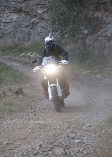 Motorradfahrer auf einer Adventure-Maschine mit eingeschaltetem Licht auf einem staubigen Schotterweg vor einer Felswand